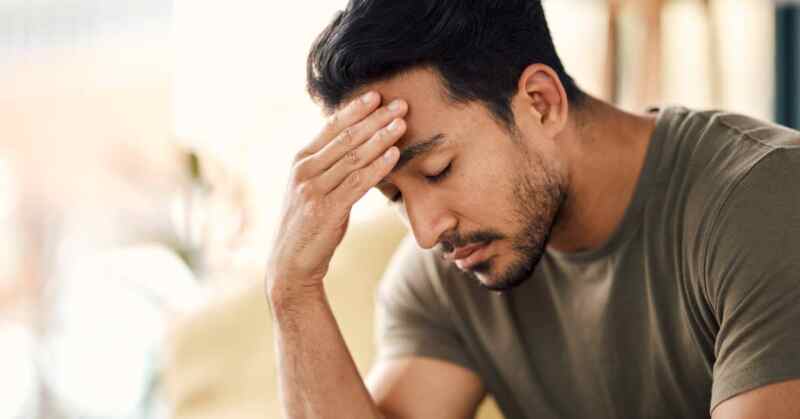 A man touches his head and appears to feel stressed. He has a beard and wears a brown and gray t-shirt.