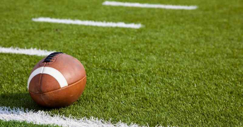 A close-up of a football lying on a green football field outside near four white lines on the grass.