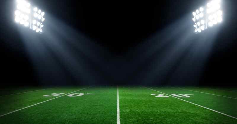 The 20 and 30-yard lines on a bright green football field are illuminated by two large stadium lights.