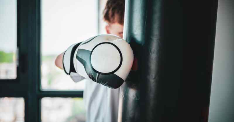 A boy wearing a boxing glove is punching a punching bag in front of a row of windows. The punching bag is in front of him.