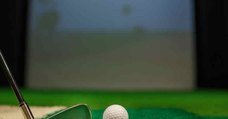 A close-up of a white golf ball resting on turf. Next to the ball is a golf club, with a simulator screen in the background.
