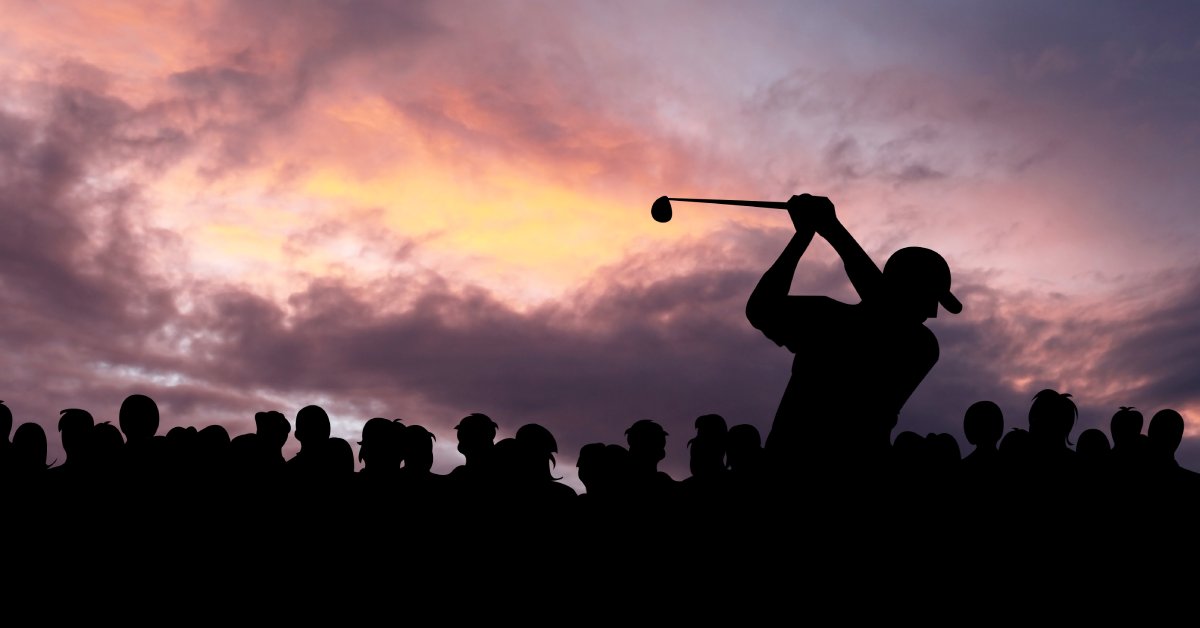 A golfer wearing a hat swings during sunset as a crowd of people looks on, behind them. Shadows obscure everyone.