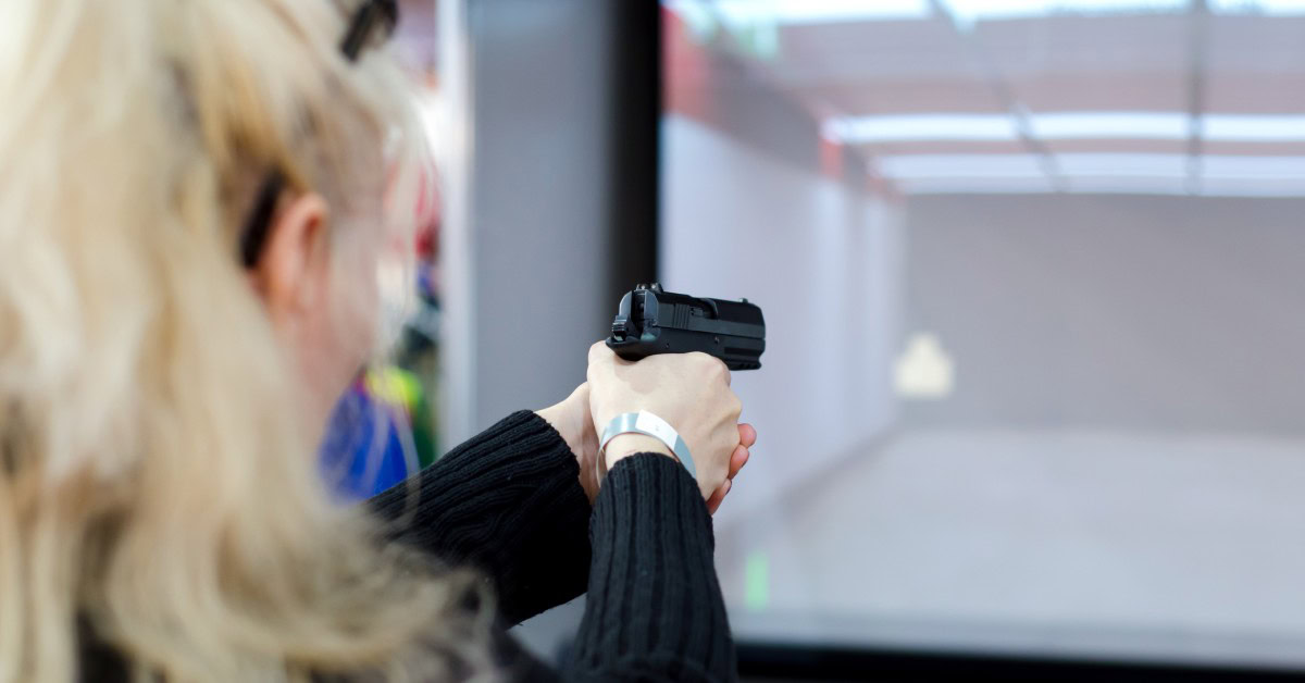 A person with blonde hair in a black top aims a handgun at a target displayed on a screen in a simulated shooting range.