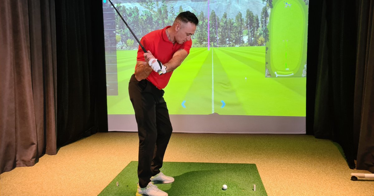 A man wearing a red polo shirt swings a golf club at a ball with his right hand in front of a simulator.