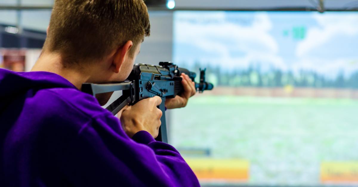 A person in a purple hoodie aims a rifle in an indoor shooting simulator with a virtual outdoor scene on a screen.
