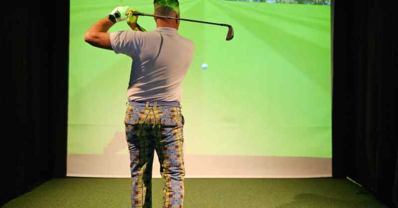 A male golfer wearing colorful pants swings a club indoors while practicing on a high-tech golf simulator screen.