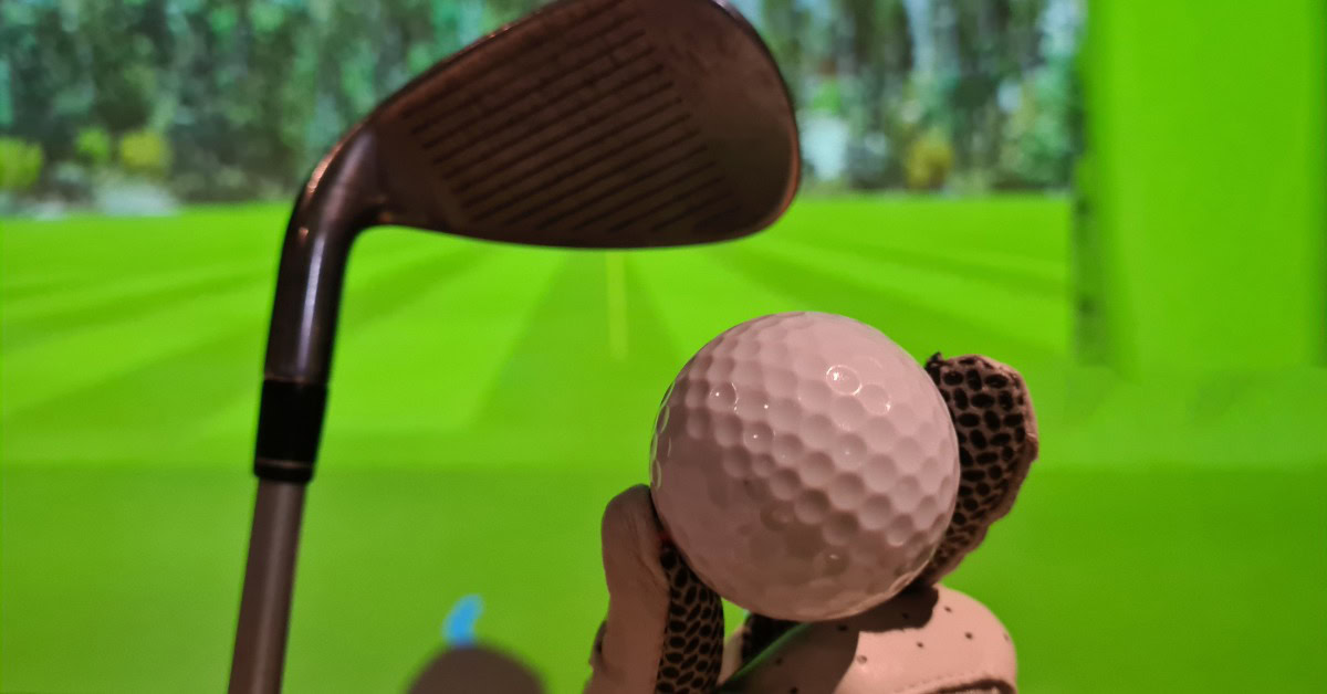 A close-up of a golf ball and club positioned in front of a bright green indoor golf simulation screen.