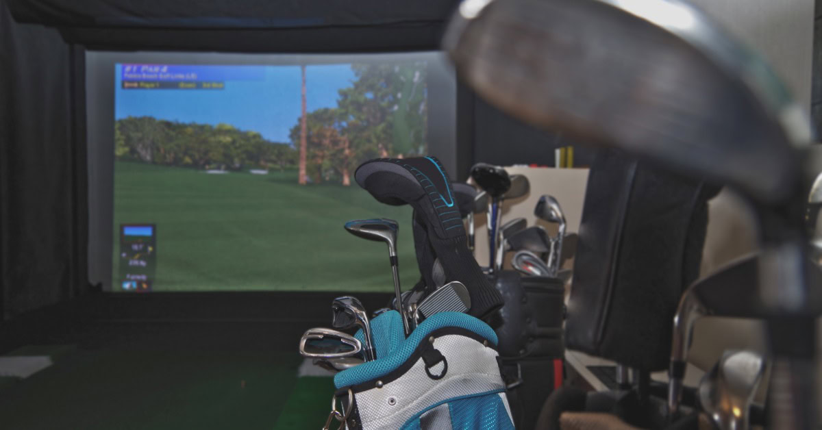 Golf clubs in a bag in the foreground of an indoor setup with a large screen displaying a virtual golf course.