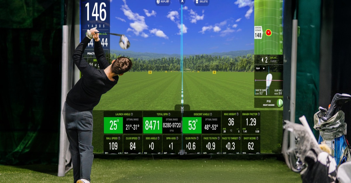 A golfer swinging in an indoor simulator with data displayed on a large screen in a high-tech training environment.
