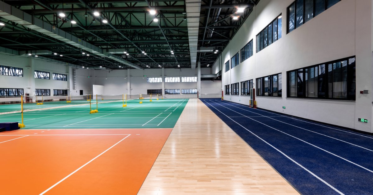 A large indoor sports complex with badminton courts, a blue running track, wood flooring, windows, and ceiling lights.