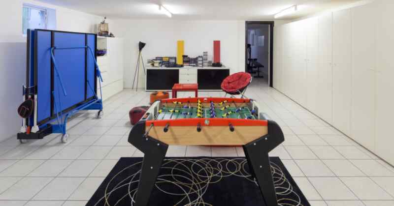 A foosball table on a patterned rug in a bright playroom with storage cabinets, a chair, and game equipment.