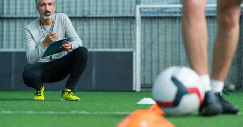 A man crouching with a clipboard on green turf near cones, a soccer ball, a standing player, and a goal.