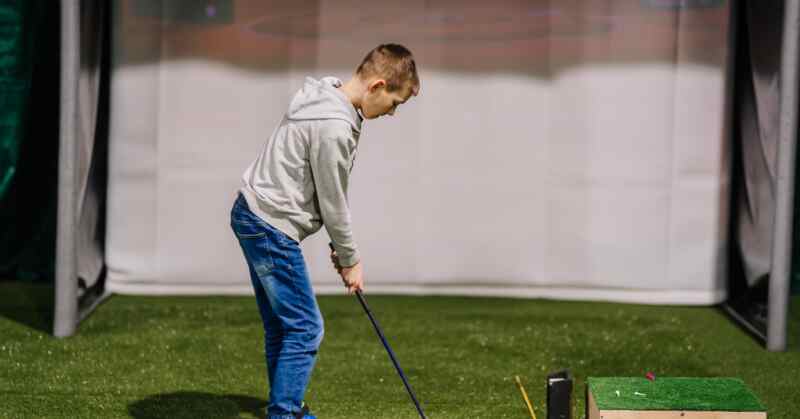 A boy in a gray hoodie standing with a golf club on green turf near golf balls and a simulator screen.