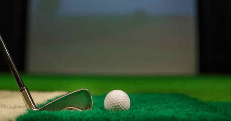 A golf ball and iron rest on artificial turf inside an indoor simulator designed for training and practice.
