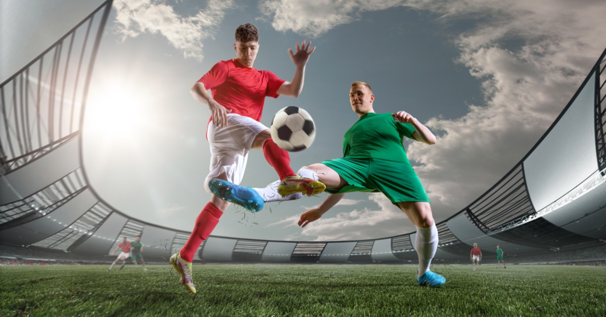 Two soccer players in red and green uniforms compete for the ball on a grass field inside a crowded outdoor stadium.