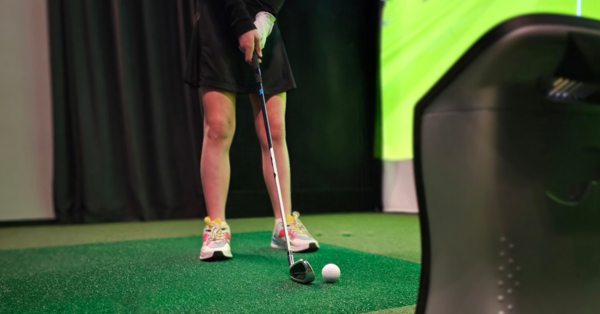 A person standing on green turf with a golf club and ball, with a black device nearby in the foreground.