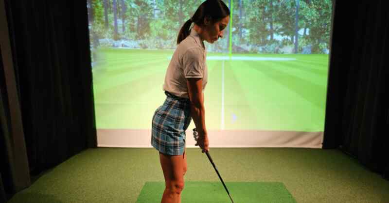 A woman holding a golf club on a green mat beside a ball, facing a projected course inside an indoor bay.