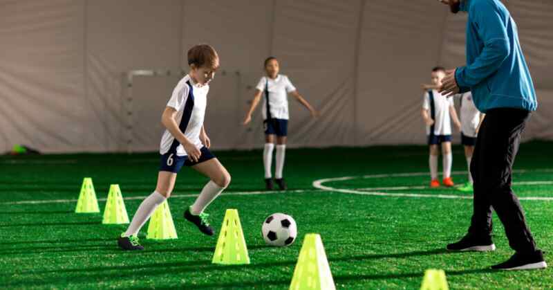 Children are practicing soccer drills with cones and a ball in front of an adult on an indoor turf field.