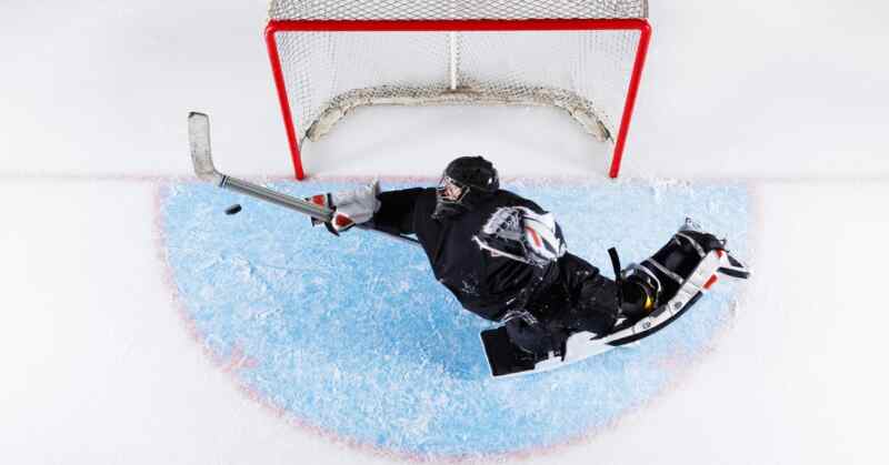 A hockey goalie in black gear reaching with a stick toward a puck in front of a red goal net on white ice.