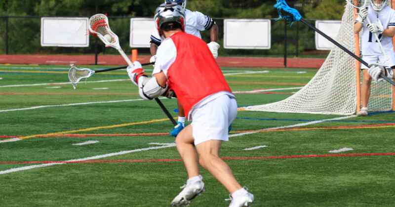 A lacrosse player wearing a red practice vest running with a stick near other players and a goal on a green turf field.