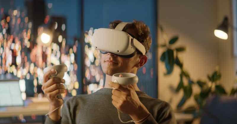 A man wearing a virtual reality headset while holding two controllers in a room with plants and window lights.