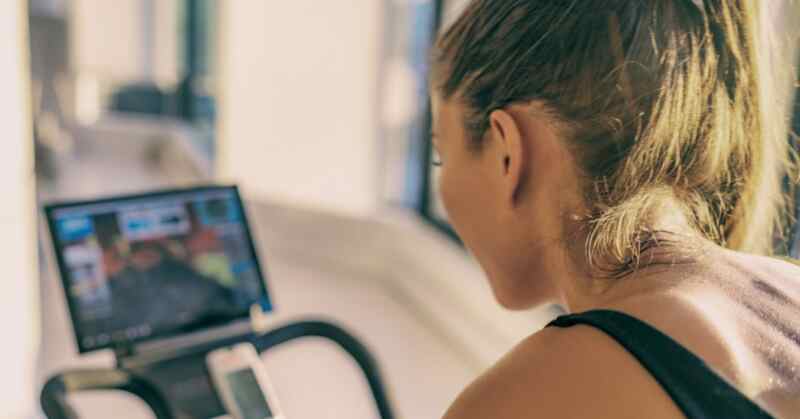 A woman riding a stationary bike indoors while facing a screen, with tied-back hair and sweat on their shoulder.