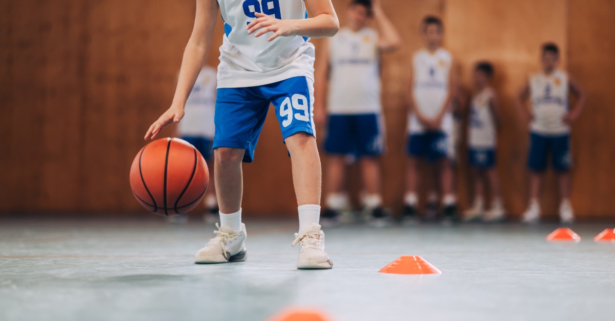 A young basketball player in a white and blue uniform dribbling a basketball between orange cones as others stand behind.