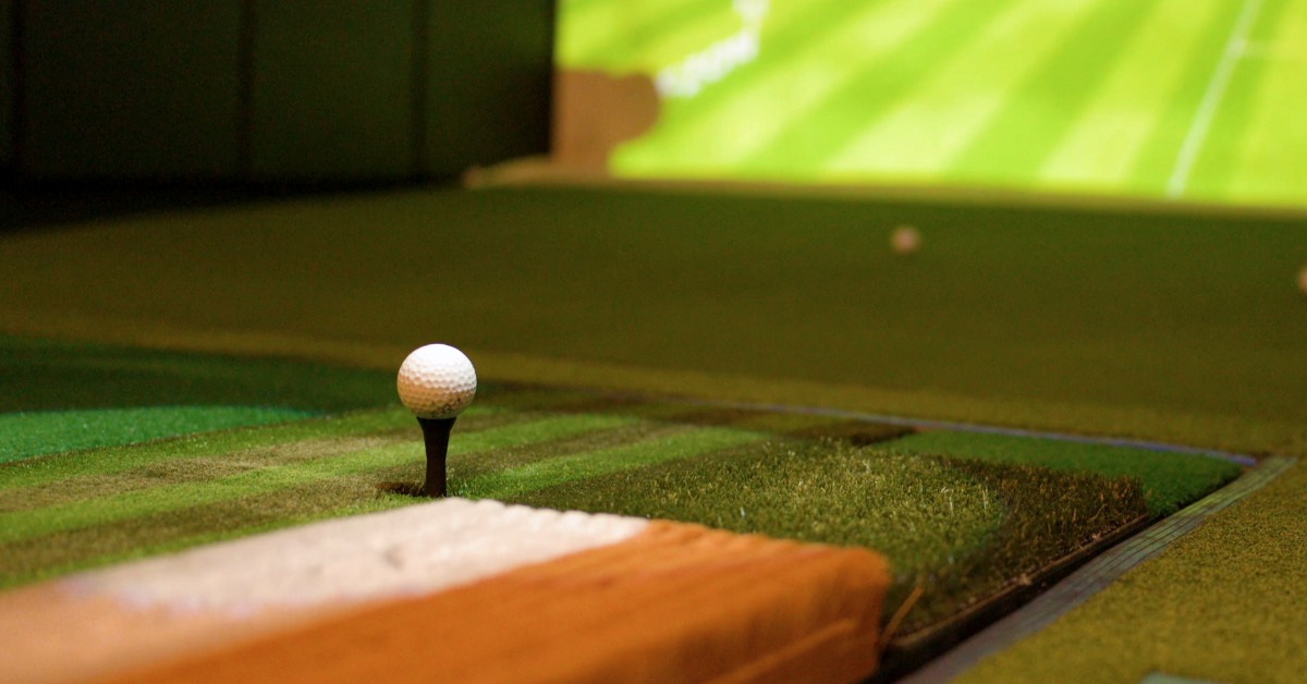 A white golf ball resting on a black tee in a patch of green artificial turf, with a large illuminated screen behind it.